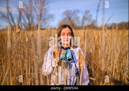 Woman looking at the camera straight on, with a neutral expression, with blue binoculars standing in a field of cat tails in a marsh during the day. Stock Photo