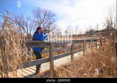A woman in a blue down jacket looks into the distance through blue binoculars on a bridge in a park on a sunny spring day. Stock Photo