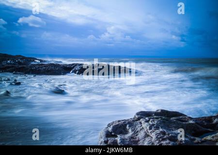 A long exposure sea waves hitting rocks with cloudy sky in the background Stock Photo