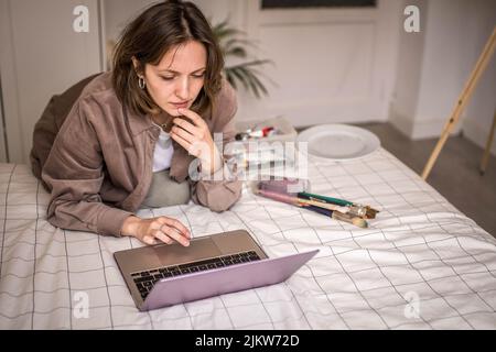 Picture of Apprentice Painter Getting Inspired Taking Ideas with the Laptop to Paint with Brushes on a Canvas with Easel in her Room Stock Photo