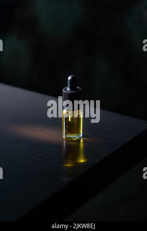A glass bottle with a pipette filled with cbd oil stands on a dark wood table, against a dark background. Stock Photo