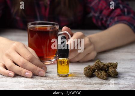 Women's hands hold a cup of tea.  in the foreground a glass bottle with a pipette with cannabis cbd oil, dry marijuana buds lie nearby Stock Photo