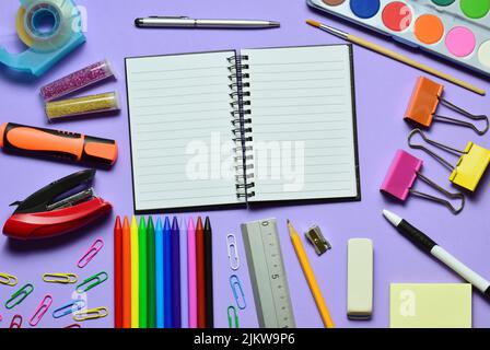 A top view of an open notebook surrounded by different stationery on mauve background Stock Photo