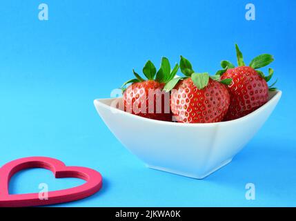 A closeup of red strawberries in a white porcelain bowl on blue background with copy space Stock Photo