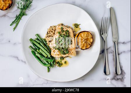 A top view of halibut fish with chimichurri sauce, green beans and lemon on a white plate on a marble table Stock Photo