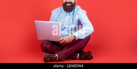 Smiling handsome bearded man worker laptop. Happy young man sitting on the floor with and using laptop computer on red background Stock Photo