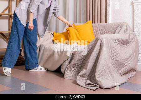 Young decorator girl lays yellow pillow on sofa covered with bedspreads. Cosily furnished living room with wooden shelf against wall. Stock Photo