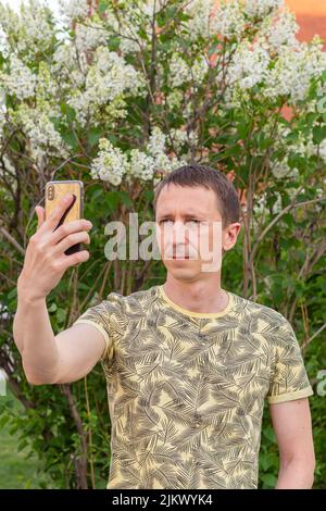 man takes selfie on his cell phone on white lilacs blooming background, close-up, side view, selective focus. Natural background and using technology Stock Photo