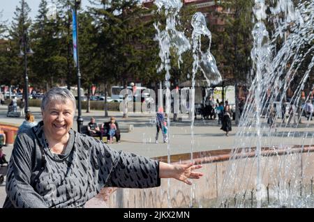 Relaxed and laughing mature senior woman sitting by fountain, seen through jets of functioning fountain with green trees and shrubs in background, enj Stock Photo