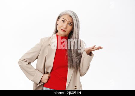 Portrait of asian senior woman, korean middle-aged woman in stylish outfit shrugging, raising empty hand up, looking questioned and confused, white Stock Photo