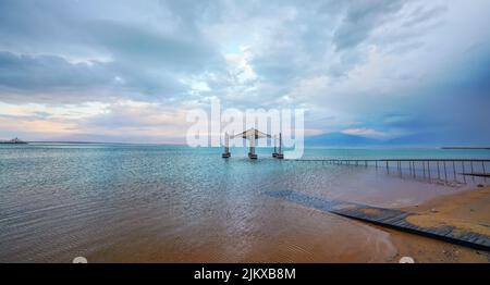 Calm morning at Ein Bokek Dead Sea beach, clouds over water surface, sun shade shelter in distance Stock Photo