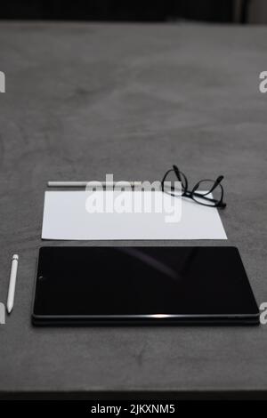 A vertical closeup of a black tablet, a piece of white paper, white pencil and a pen, and eyeglasses Stock Photo