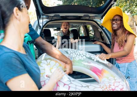 Multiethnic family and friends loading bags in car, preparing baggage in automobile. Travelling to seaside with diverse people, putting luggage and inflatable in vehicle, leaving on vacation. Stock Photo