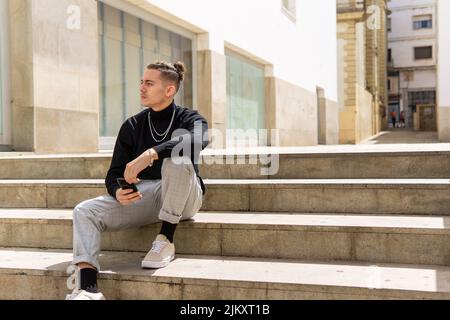 Portrait of a young mixed-race man thoughtful holding a smartphone sitting on stairs in the city. Social media, communication, mobile apps, business, Stock Photo