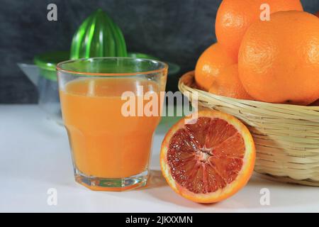 Glass with orange juice on white table with basket full of oranges and citrus squeezer next to it Stock Photo