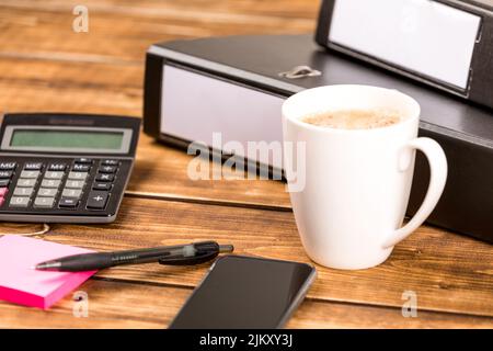 A selective shot of a cup of coffe with calculators folders and different supplies on a wooden office desk Stock Photo