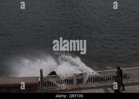 A high angle view of two people walking by the pier near the water crashing on the rocks Stock Photo