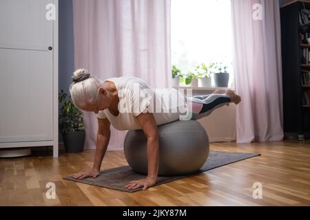 Sporty senior woman doing yoga plank using a ball exercising in living room Stock Photo