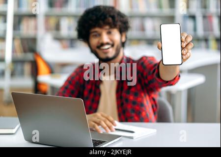 Mock-up and copy-space concept. Photo of defocused Indian or Arabian male student, sitting at table in library, showing his smart phone with blank white screen for advertising and presentation, smile Stock Photo