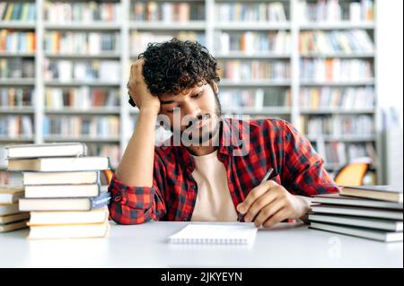 Back to school. Indian or Arabian bored tired male student, sitting at the table with books in the university library, takes notes in a notebook, looking sad and exhausted from studying, needs a break Stock Photo