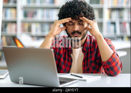 Tired tense indian or arabian guy, university student, wearing stylish shirt, sits at a table in the library, closed his eyes, massages his whiskey, tired of boring studies, has a headache,needs break Stock Photo