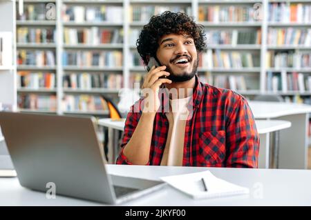 Joyful handsome positive curly haired stylish mixed race guy, student, sits at a table in the library, has a pleasant phone conversation with a friend, looks away, smiling friendly Stock Photo