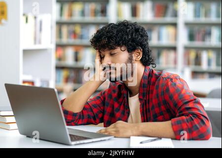 Chronic sleep deprivation. Tired bored arabian or indian university student, is sleeping while sitting at desk in library, exhausted of learning, preparing for exam session, suffering of lack of sleep Stock Photo