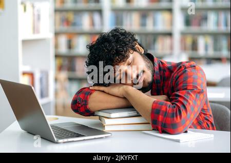 Tired bored arabian or indian university student, is sleeping while sitting at a desk in a library, exhausted of learning, preparing for exam session, suffering of stress and chronic sleep deprivation Stock Photo
