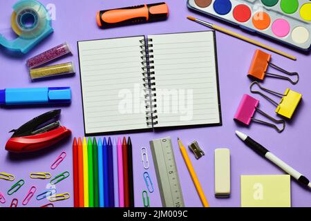 A top view of an open notebook surrounded by different stationery on mauve background Stock Photo