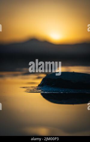 A vertical shallow focus of a rock reflecting on shiny yellow water with blurred sunset cloudy sky Stock Photo