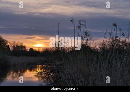 A warm summer sunset shining through the clouds with its reflecting in a lake surrounded by greenery Stock Photo