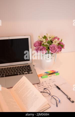 A vertical shot of a decorative vase with pink roses on a desk with stationery and a laptop Stock Photo