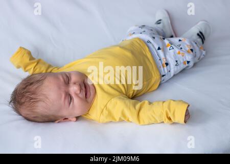 A closeup of a Caucasian baby boy, wearing a yellow shirt, lying on a white fabric Stock Photo