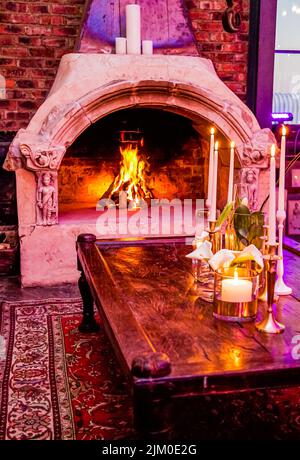 Beautiful candles in candlesticks on the old-style wooden table in a cozy living room with a fireplace Stock Photo