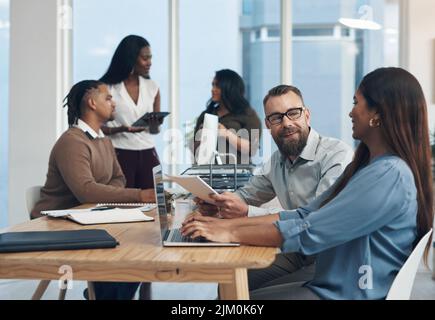 Can I get your help on this task. two young businesspeople sitting together and using a laptop while their colleagues work in the background. Stock Photo