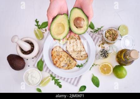 Avocado and cream cheese toasts preparation - Woman holding halved avocado over table with ingredients Stock Photo