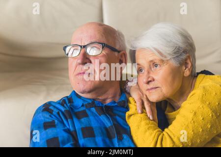 Relaxed loving married elderly man and woman hugging and ooking away, wife resting on her husband's shoulder while sitting on the floor near the couch in the living room at home. High quality photo Stock Photo