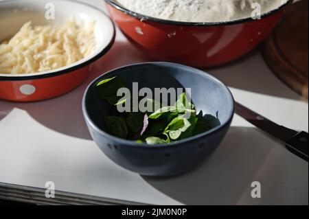 Close-up. Freshly picked basil leaves in navy bowl, nearby vintage bowls with grated cheese and yeast dough on a table Stock Photo