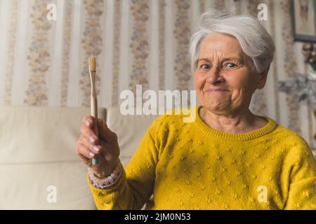 Funny caucasian elderly woman, sitting on a beige couch in the living room at home and holding a toothbrush in one hand, looking shocked and excited at the same time. High quality photo Stock Photo