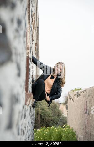 A vertical selective focus shot of a Caucasian woman hanging from a building's wall, smiling Stock Photo