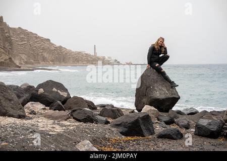 A Caucasian young woman sitting on a rock by the Aegean sea, Santorini Stock Photo