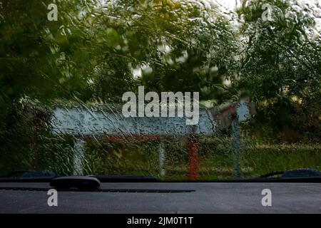 A closeup of a rainy car windshield looking out into a park with a Bluetooth earpiece on the dashboard Stock Photo