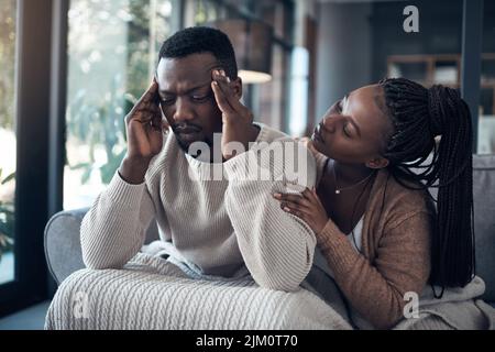 Do you need me to get you anything. a handsome young man sitting and suffering from a headache while his girlfriend comforts him at home. Stock Photo