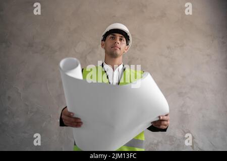 Young arabic architect man with helmet and holding blueprints isolated on grey background, construction engineer. Builder with drawings. Stock Photo