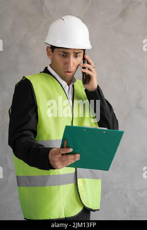 Angry muslim engineer or architect screaming while talking on the phone, builder holding clipboard and cursing into smartphone, isolated on gray Stock Photo