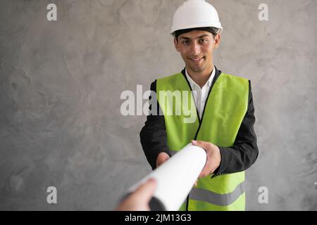 Young arabic architect man with helmet and holding blueprints isolated on grey background, construction engineer in a hard hat. Builder with drawings. Stock Photo