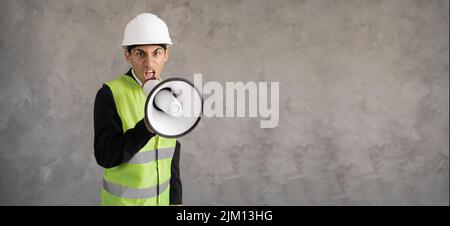 Arabic male contractor in hardhat standing with megaphone on gray background and shouting with loud and noise, engineer or foreman shouting into a Stock Photo