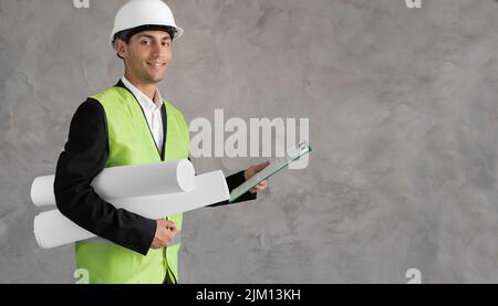 Young arabic civil engineer holding paperwork blueprint roll standing post in studio. Content male construction architect in suit and hardhat with Stock Photo