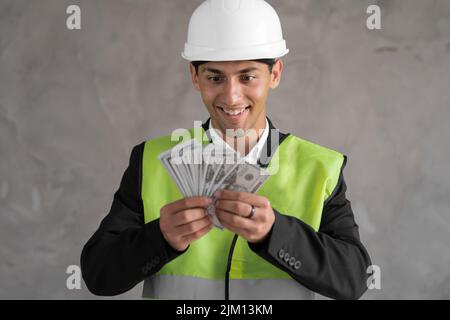 Young arabic builder man engineer wearing safety helmet holding dollars as payment over background with raised hand, payment construction Stock Photo