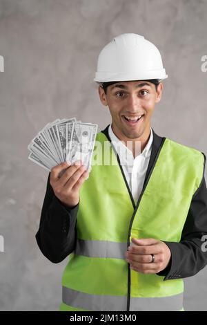 Young arabic builder man engineer wearing safety helmet holding money dollars as payment over background with raised hand, payment work construction Stock Photo
